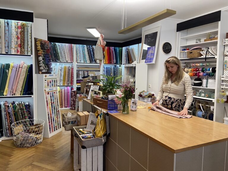 Eine Mitarbeiterin schneidet an einem großen Holztisch rosa Stoff zu; im Hintergrund sieht man Regale voller bunter Stoffballen und Nähzubehör.