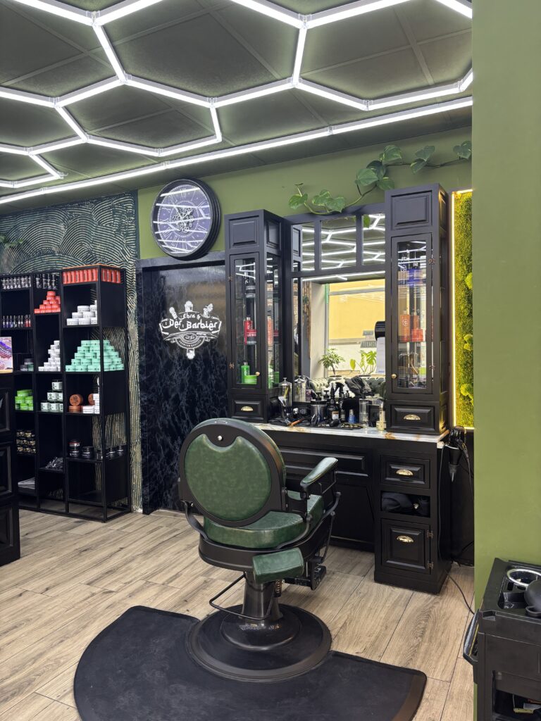 Ein klassischer grüner Leder-Barbiersessel vor einem großen Spiegel und einem schwarzen Holz-Frisiertisch mit goldenen Akzenten.
