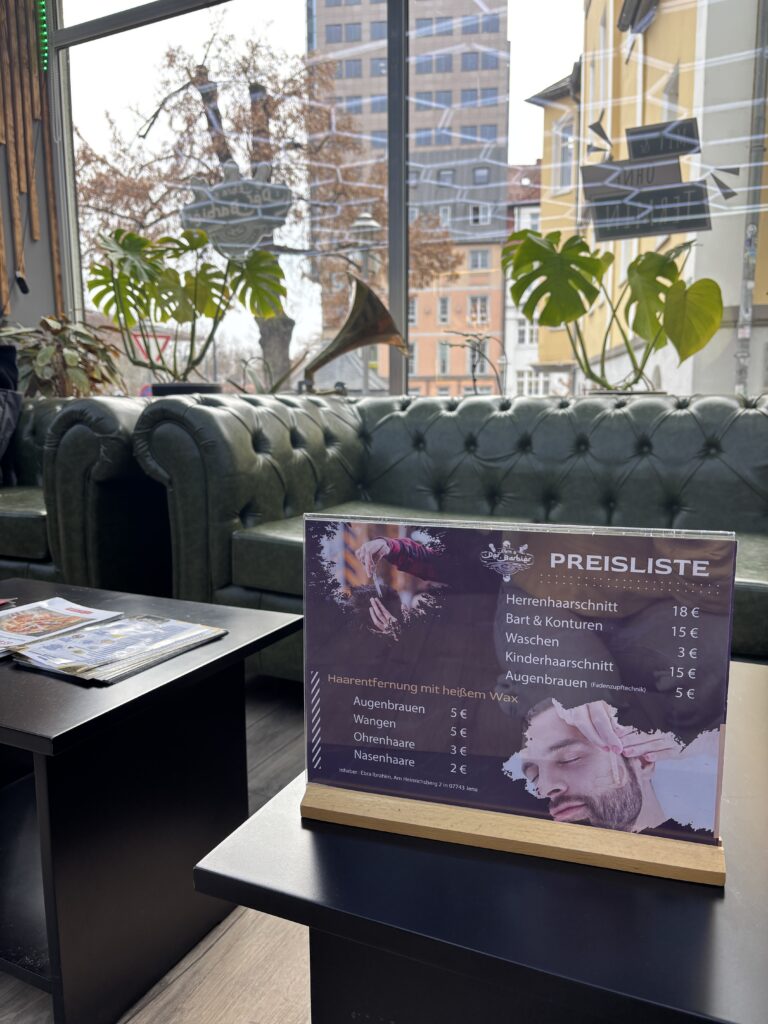 Eine Preisliste auf einem Tisch vor einem dunkelgrünen Chesterfield-Sofa und einem großen Fenster mit Blick auf Jena.