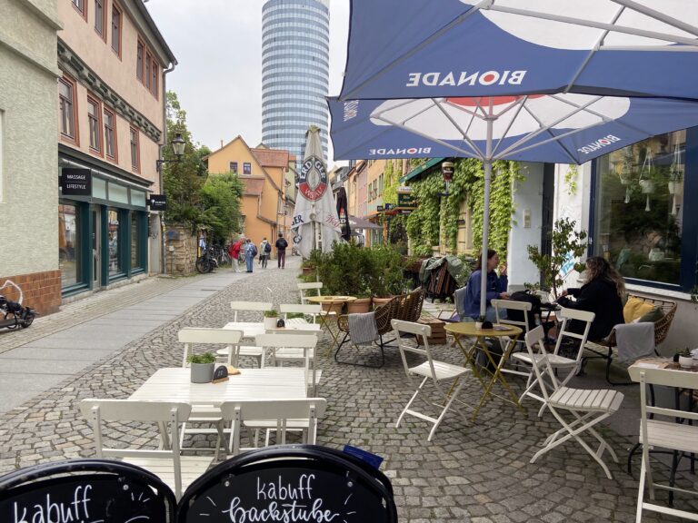 Außenbereich des Cafés in der gepflasterten Wagnergasse mit weißen Tischen und Stühlen; im Hintergrund ragt der Jentower in den Himmel.