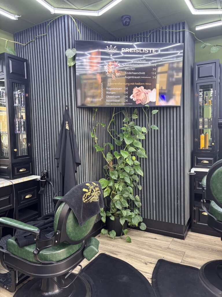 Ein Flachbildschirm an einer dunkel gestreiften Wand zeigt die Preisliste des Barbiers neben einem grünen Sessel.