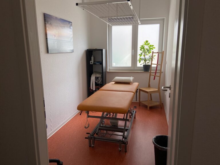 Eine Behandlungsliege steht in einem hellen Therapieraum mit Fenster, Pflanze und weiteren Ausstattungsgegenständen.