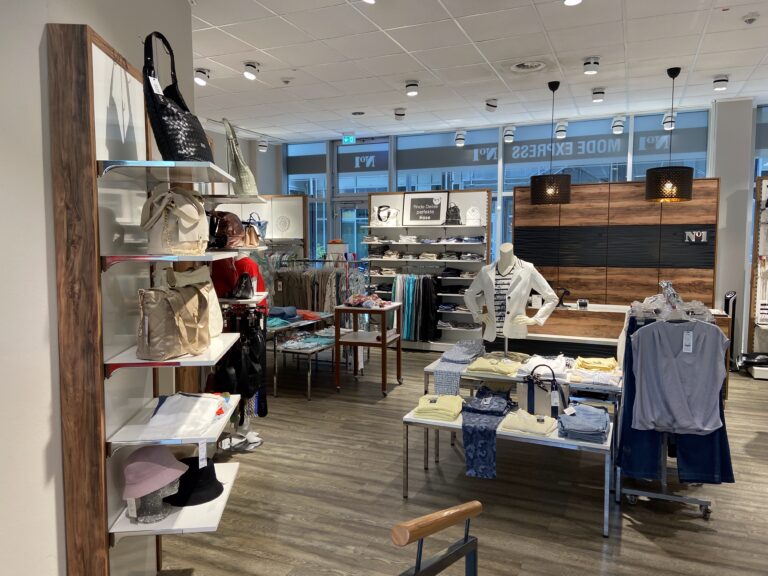 Blick in den weitläufigen, hell erleuchteten Verkaufsraum mit Kleiderständern, Tischen mit gefalteter Ware und Regalen mit Handtaschen.