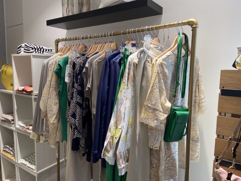 Nahaufnahme einer Kleiderstange mit farblich sortierter Damenmode in Grün-, Beige- und Blautönen; eine grüne Handtasche hängt dekorativ an der Stange.