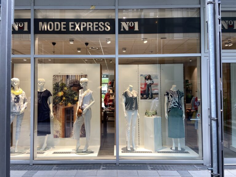 Außenansicht der großen Glasfront von Mode Express No1 mit vier Schaufensterpuppen, die aktuelle Outfits in Blau, Weiß und Grün präsentieren.