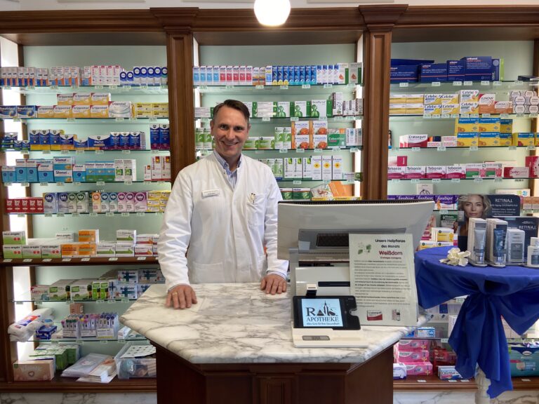 Der Apotheker steht freundlich lächelnd im weißen Kittel hinter einem edlen Marmor-Handverkaufstisch; im Hintergrund sieht man klassische Holzregale mit Medikamenten und einen Bildschirm mit dem Apotheken-Logo.