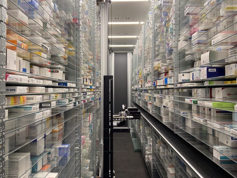 Blick in das Innere eines hochmodernen, automatischen Warenlagers (Kommissionierautomat) mit Greifarm und unzähligen Medikamentenpackungen in geordneten Reihen.