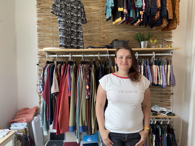 Die Inhaberin steht freundlich lächelnd in einem weißen T-Shirt mit dem goldenen Aufdruck "FRAENNE" vor einem Kleiderständer und einer Wandverkleidung aus Bambus.