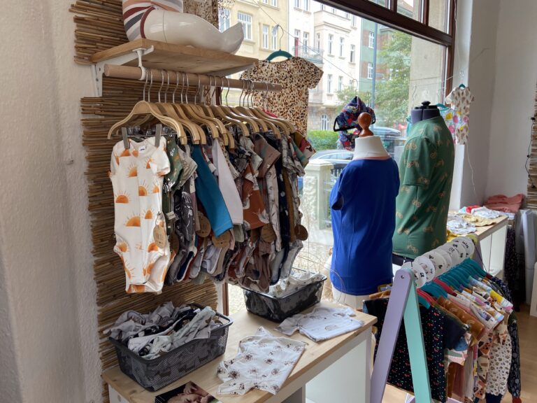 Kleiderständer am Schaufenster gefüllt mit bunter Baby- und Kinderkleidung, darunter Bodys mit Sonnenmuster und Shirts mit Fahrradmotiven