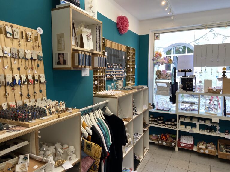 Blick in den Verkaufsraum mit markanten türkisblauen Wänden, weißen Regalen voller Accessoires, Kleiderstangen und einem großen Schaufenster zur Straße.