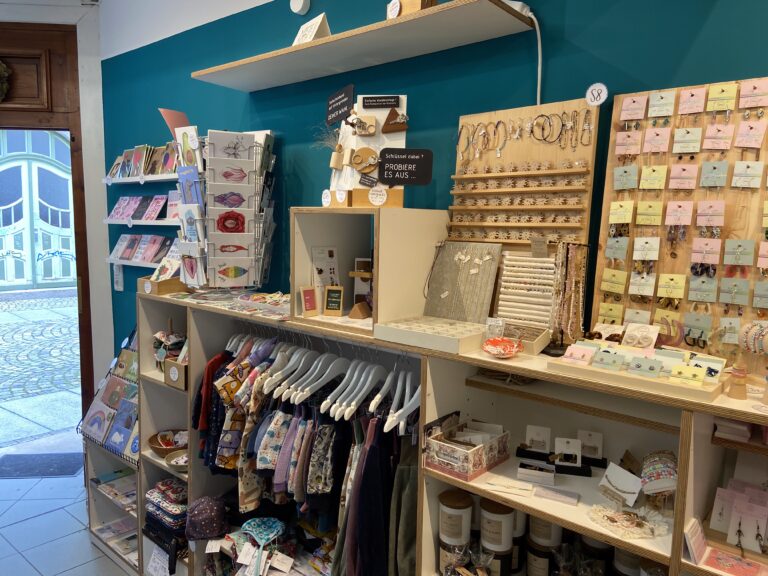 Eingangsbereich mit geöffneter Tür, einem Ständer voller Postkarten, bunter Kinderkleidung auf Bügeln und Regalen mit Geschenkideen.