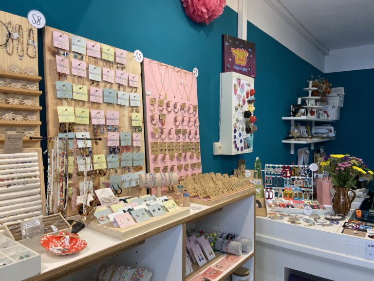 Der Verkaufstresen, dekoriert mit frischen Blumen, ist umgeben von Schmuckständern, Armbändern, Stickern und vielen kleinen Accessoires.
