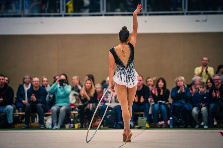 Rückenansicht einer Rhythmischen Sportgymnastin im Glitzer-Trikot mit einem Reifen, die vor einem applaudierenden Publikum in der Halle steht.