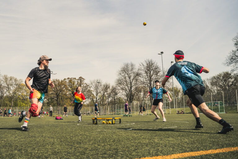 Junge Menschen spielen bei sonnigem Wetter Roundnet (Spikeball) auf einem Kunstrasenplatz, ein Spieler hechtet dem Ball hinterher.