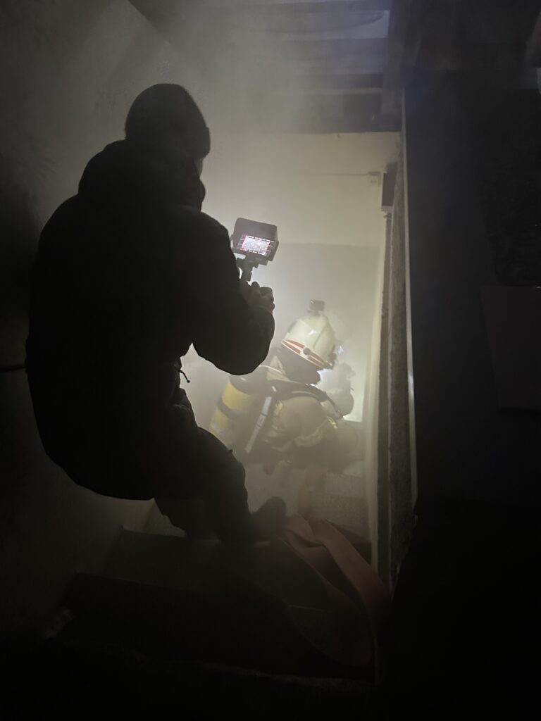 Ein Kameramann filmt in einem vernebelten Treppenhaus einen Feuerwehrmann in voller Montur während eines Einsatzes oder einer Übung.