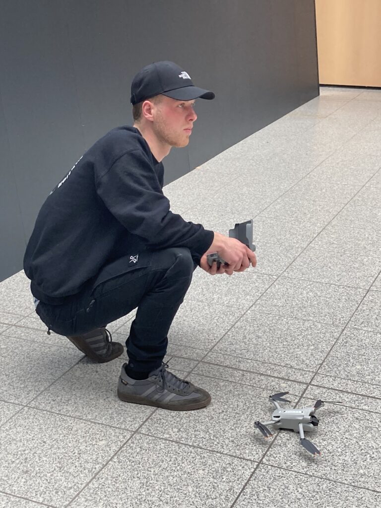Ein Videograf kniet konzentriert mit einer Fernsteuerung am Boden, vor ihm steht eine Drohne bereit zum Abflug.