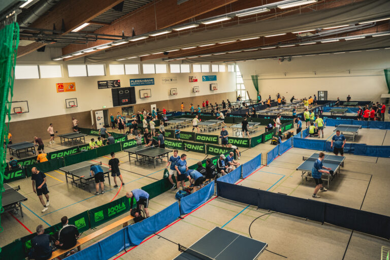 Große Sporthalle voller Tischtennisplatten, an denen zahlreiche Spieler während eines Turniers aktiv sind; Zuschauer sitzen an den Seiten.