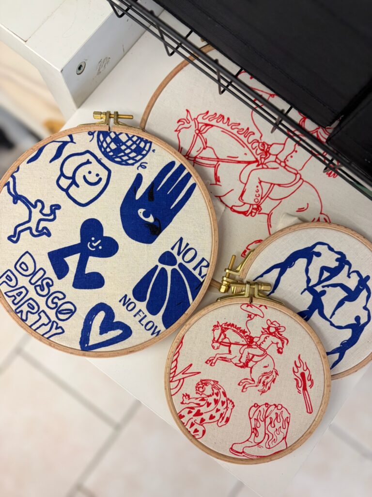 Mehrere runde Holz-Stickrahmen mit verschiedenen blau und rot illustrierten Motiven wie „Disco Party“ oder Cowboy-Illustrationen.