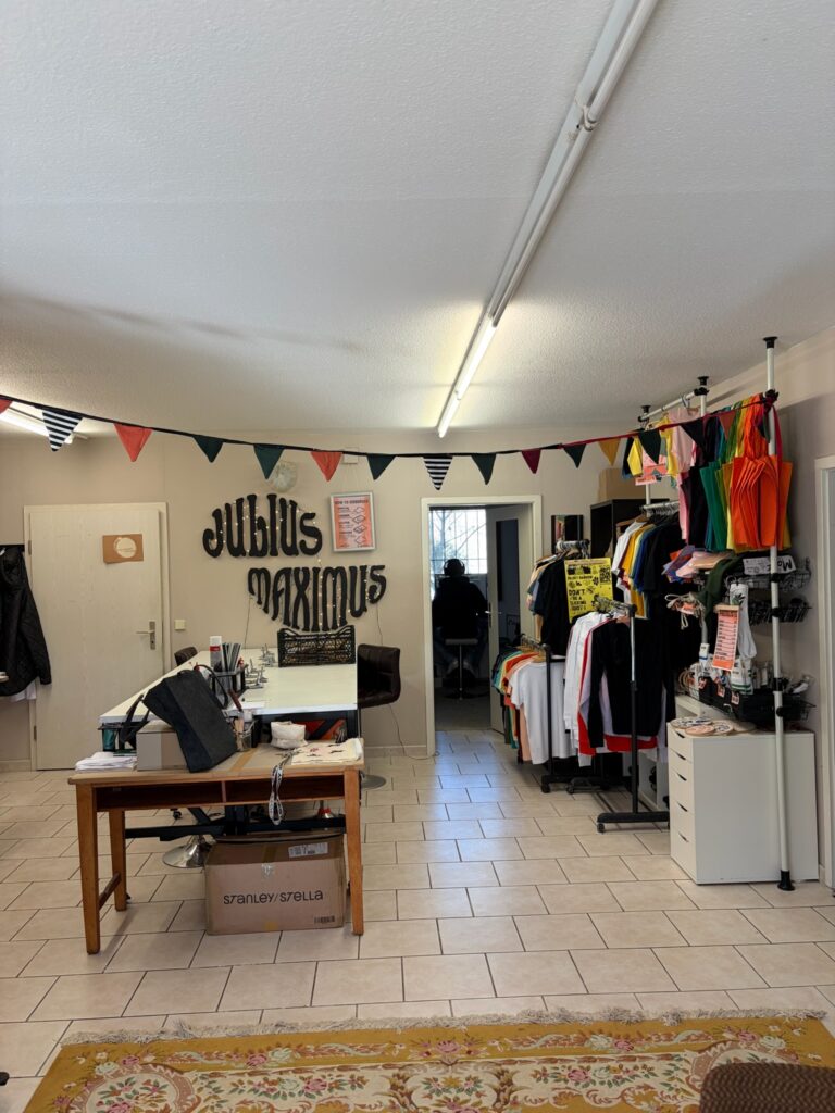 Blick in die helle Werkstatt mit großen Arbeitstischen, bunten Wimpelketten und Regalen voll farbenfroher Textilien.