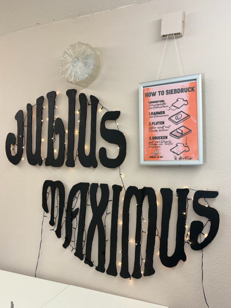 Schwarzer Schriftzug „Julius Maximus“ an der Wand neben einer gerahmten Anleitung mit dem Titel „How to Siebdruck“