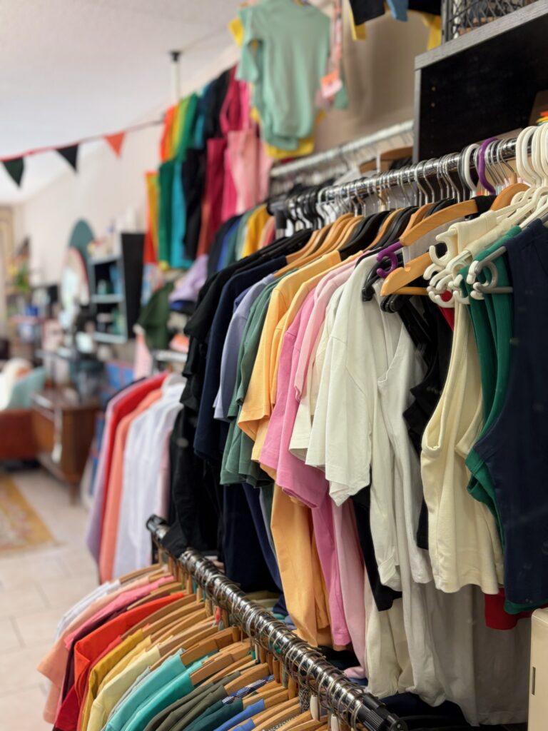Eine lange Kleiderstange voll mit bunten T-Shirts und Oberteilen in Pastell- und Leuchtfarben.