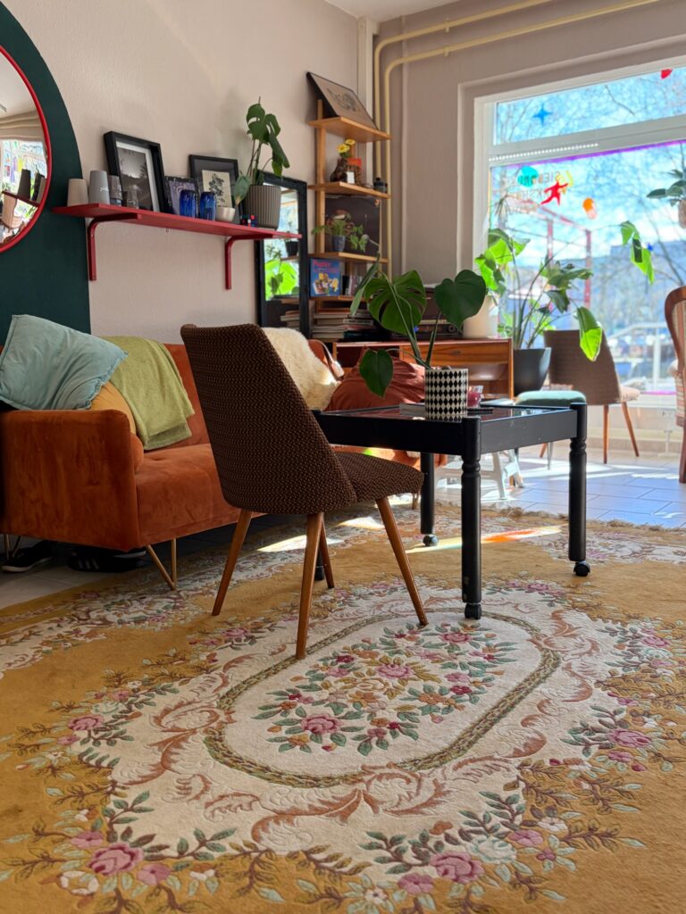 Einladende Sitzecke mit einem prachtvoll gemusterten Teppich, Vintage-Möbeln, Pflanzen und einem großen Fenster im Ladenbereich.