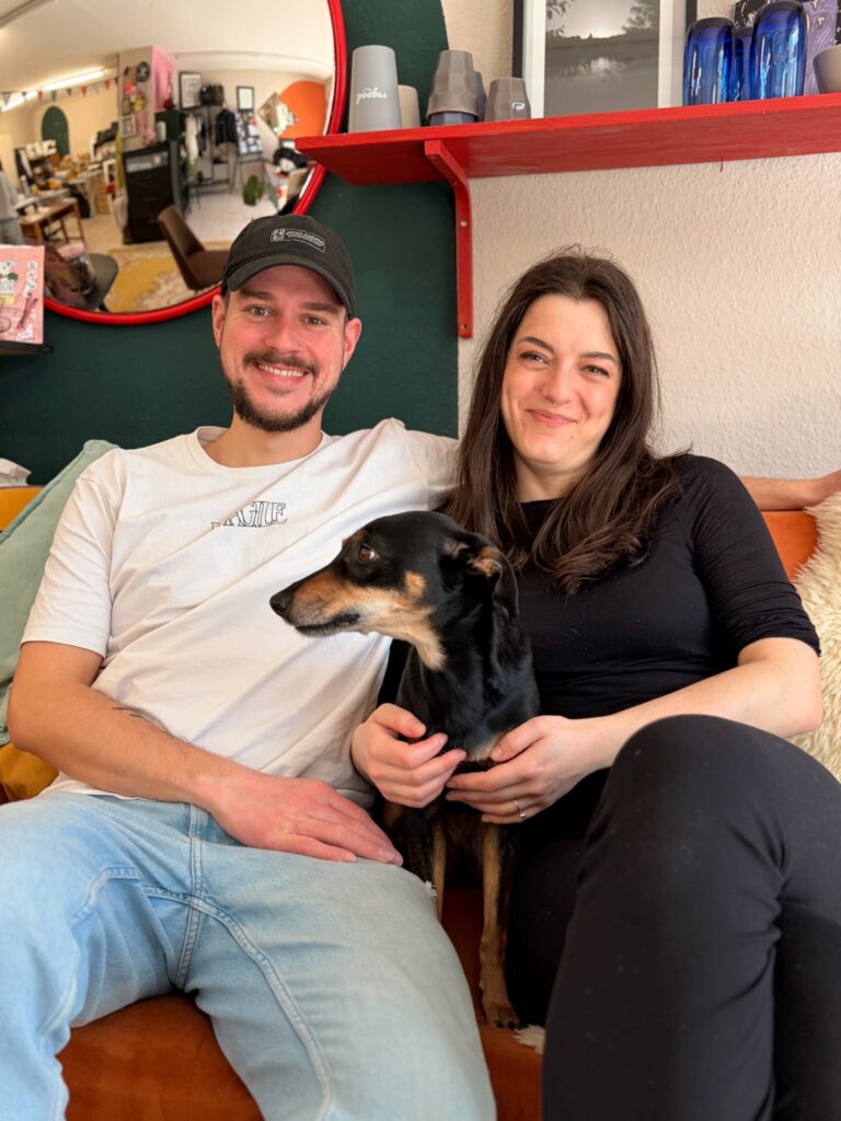 Jules und ihr Freund Phillipp sitzen lächelnd mit ihrem schwarzen Hund auf einem orangefarbenen Sofa im Ladenbereich.