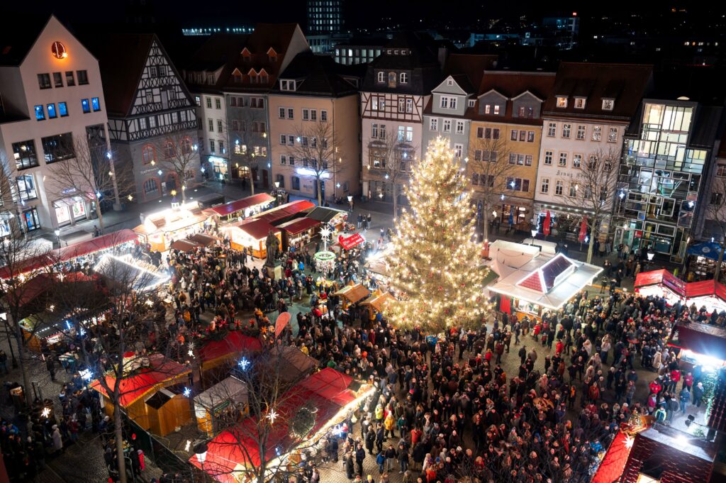 Jenaer Weihnachtsmarkt aus der Vogelperspektive mit großem beleuchteten Weihnachtsbaum, Marktständen und vielen Besucherinnen und Besuchern auf dem Marktplatz.