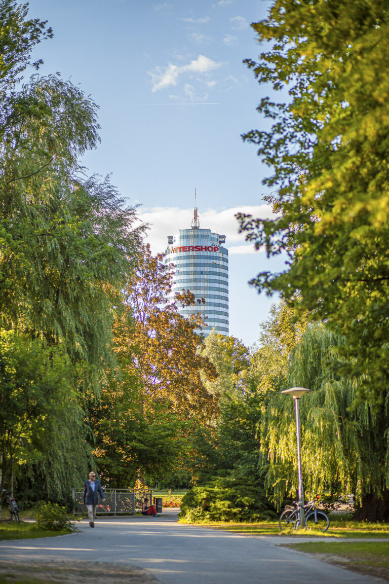 Blick durch grüne Bäume auf den JenTower unter blauem Himmel in Jena.