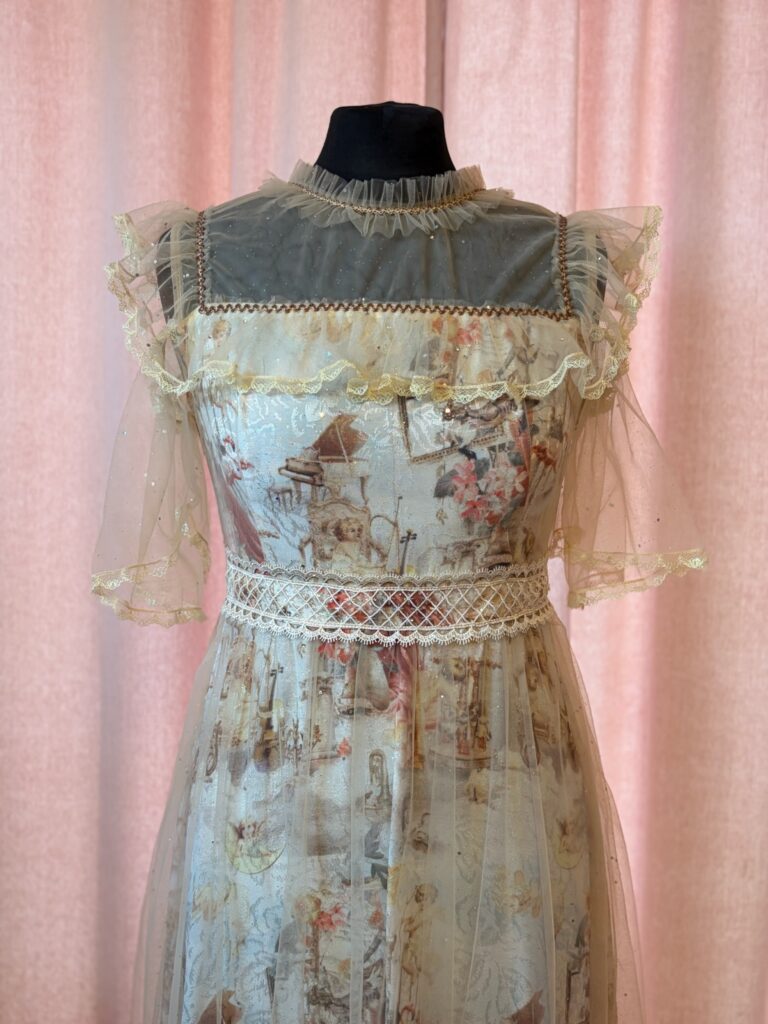 Ein helles Lolita-Kleid mit aufwendigem Print, Spitzenkragen und transparenten Ärmeln auf einer schwarzen Schneiderpuppe vor rosa Hintergrund.
