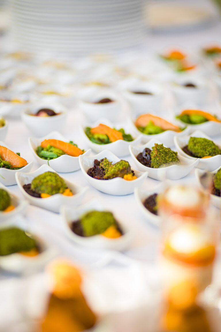 Viele kleine weiße Schälchen mit angerichtetem Fingerfood, unter anderem Lachs und grüne Toppings, auf einer weißen Tafel.