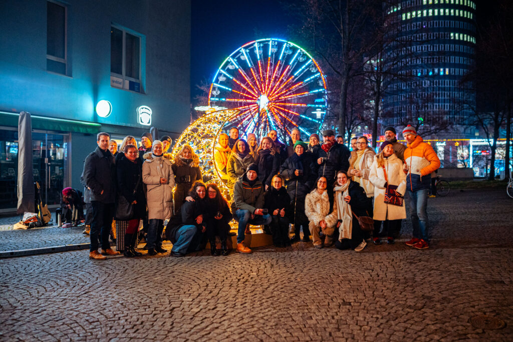 Gruppenfoto bei der Einweihung des leuchtenden Sichelmondes im Dezember 2025 während des After-Work-Advents in der Jenaer Innenstadt. Menschen stehen um den erleuchteten Sichelmond herum. Im Hintergrund sieht man ein Riesenrad.