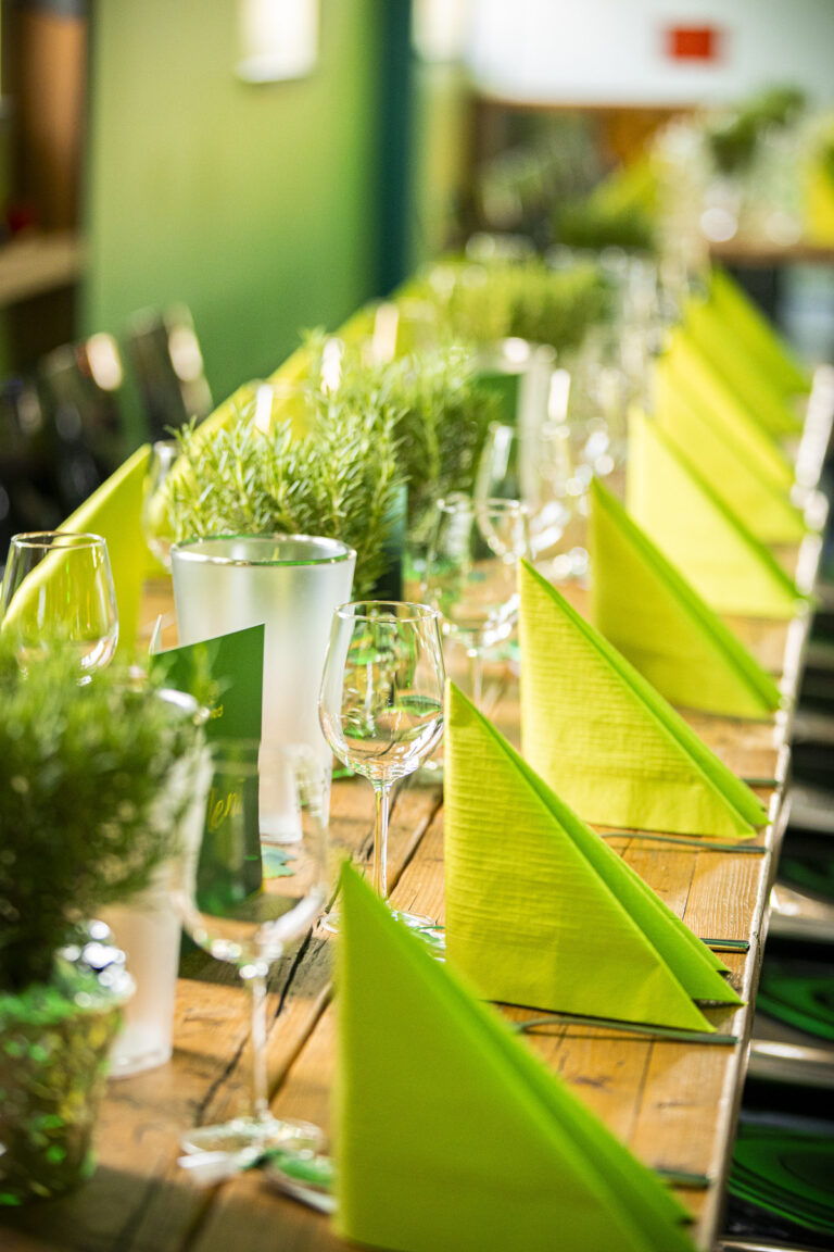 Eine lange hölzerne Tafel, festlich gedeckt mit hellgrünen Servietten, Weingläsern und frischen Kräuterpflanzen als Dekoration.