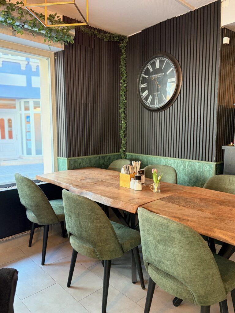 Holztisch mit grünen Stühlen steht in einem Restaurant neben einer Wand mit Uhr und Pflanzen