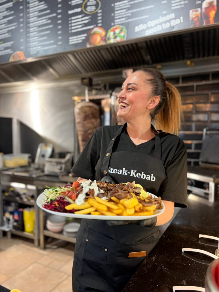 Die Inhaberin Diana steht in ihren Restaurant hinter dem Tresen und hält einen Steakteller mit Pommes, Salat und Fleisch